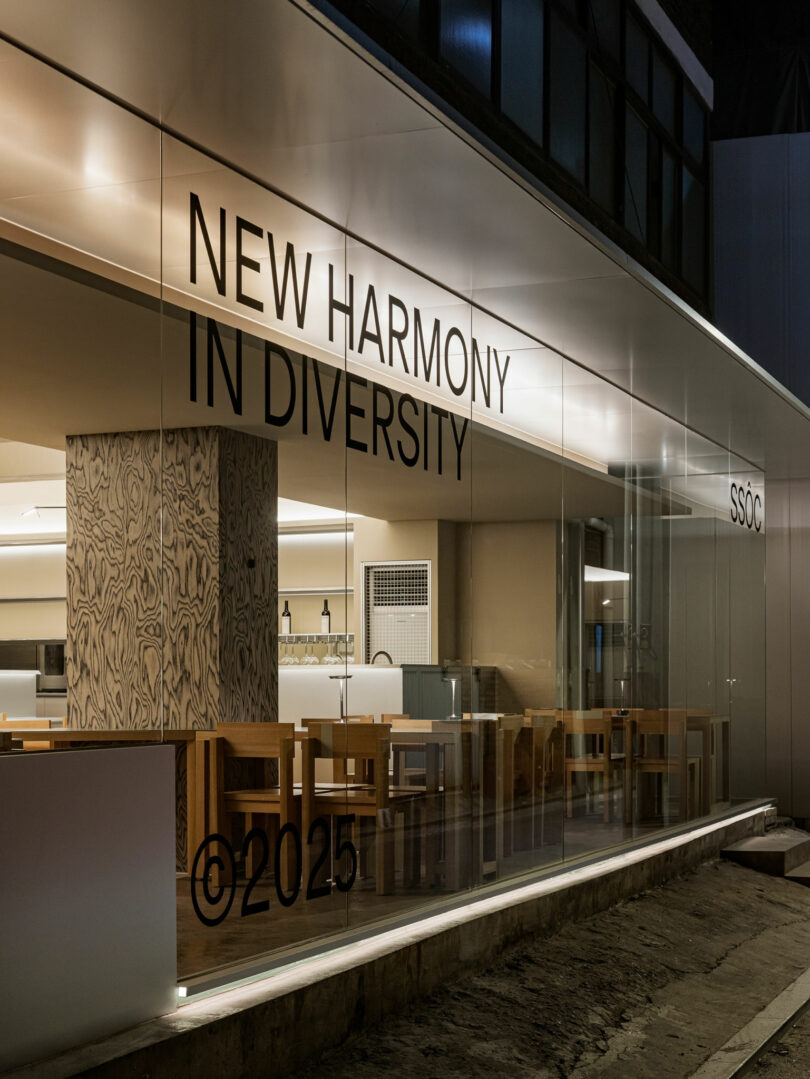 Modern interior of a building with wooden tables and chairs, seen through glass walls displaying the text "NEW HARMONY IN DIVERSITY ©2025 SSOC.