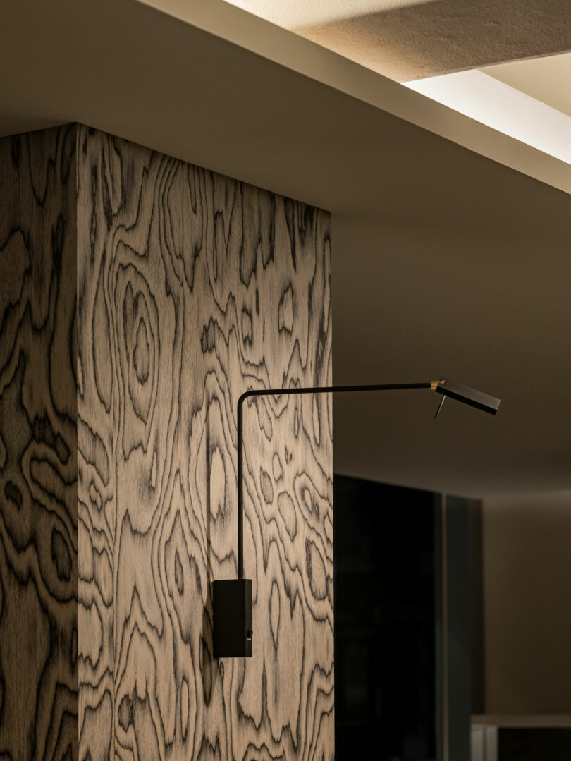 A modern black wall-mounted reading lamp is attached to a wall with a light wood grain pattern, illuminated by soft ceiling lighting.