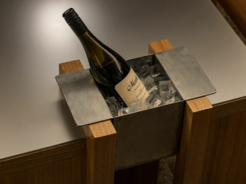 A bottle of white wine sits in a metal ice bucket filled with ice cubes, built into a wooden table.