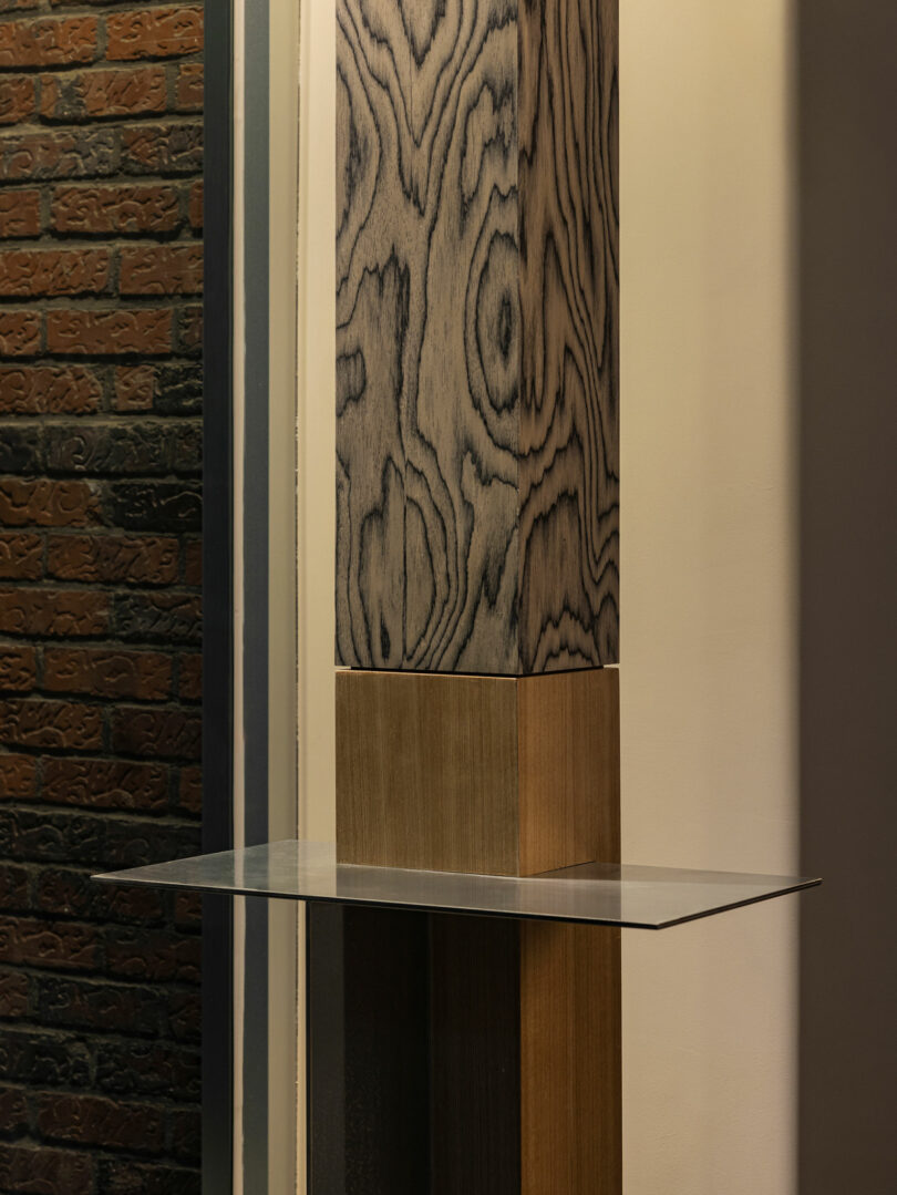 A vertical column with a wood grain pattern stands next to a brick wall; a small, flat, rectangular metal shelf extends from the column.