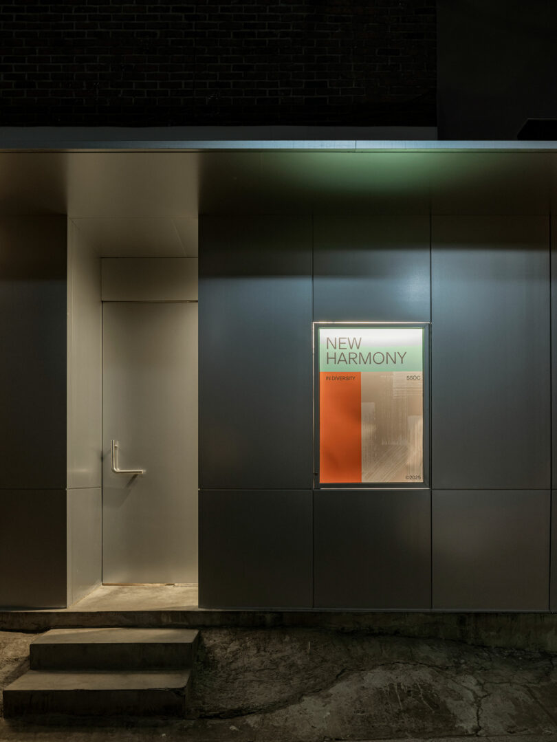 A modern gray exterior wall with a single closed door and a poster in a lit window reading “NEW HARMONY” at night. Concrete steps lead up to the entrance.