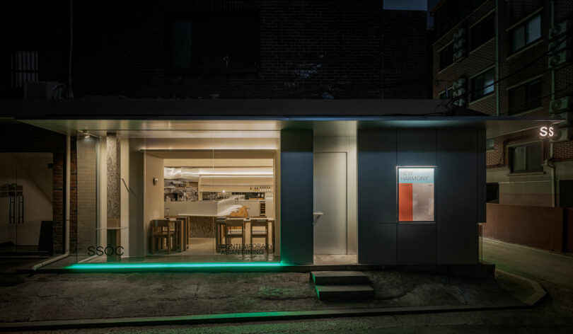 A modern storefront with large windows, minimalist signage, and interior lighting highlighting a bar and shelves inside. The exterior features clean lines and subtle green lighting.