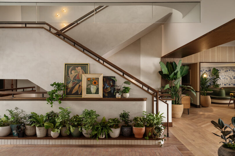 A modern interior at Roost Rainey features potted plants lined along a staircase railing, two framed paintings resting on the ledge, and more greenery and cozy seating visible in the background.
