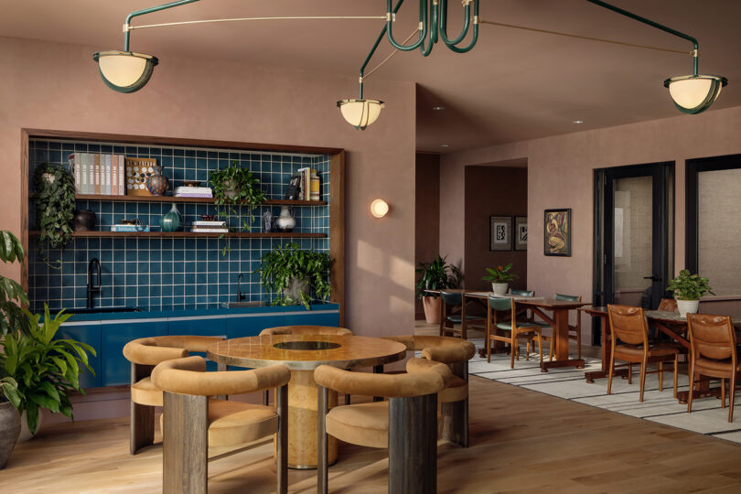 Modern lounge at Roost Rainey with a round wooden table, cushioned chairs, blue-tiled kitchenette featuring open shelving and plants, plus an adjacent dining area with leather chairs.