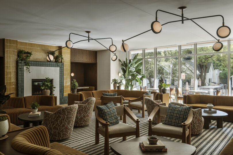 A modern lounge at Roost Rainey features patterned armchairs, a striped rug, round tables, indoor plants, and large windows overlooking outdoor greenery. Contemporary lighting fixtures hang from the ceiling.