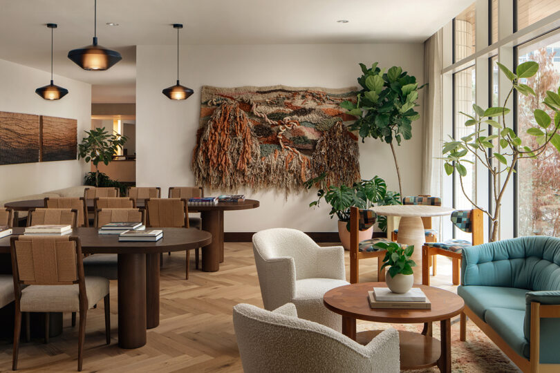 A modern lounge at Roost Rainey features wooden tables, upholstered chairs, potted plants, and large windows. A textured wall hanging decorates the white wall while books and greenery adorn the tables.