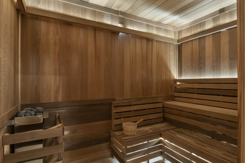 A wooden sauna room with tiered bench seating by Roost Rainey, featuring a bucket with a ladle on the bench and a heater filled with stones in the corner.