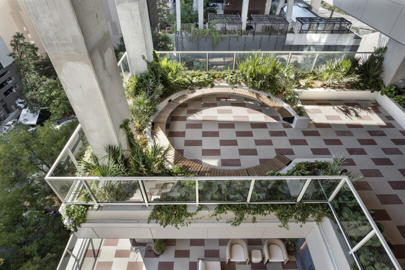 A rooftop terrace at Roost Rainey features checkered tile flooring, a circular wooden bench, planters with greenery, and glass railings overlooking the cityscape.