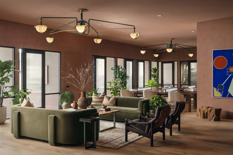 Modern lounge area at Roost Rainey with green sofas, armchairs, indoor plants, large windows, contemporary lighting fixtures, and abstract wall art.