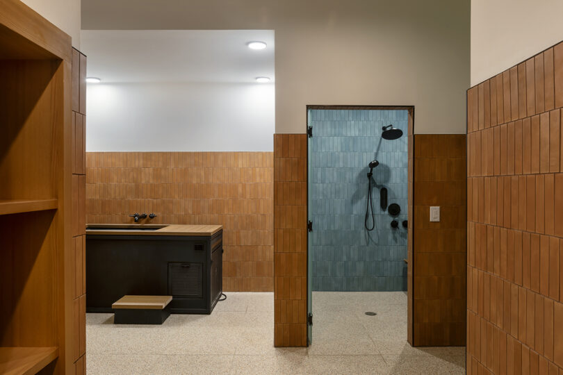 A tiled room with wood accents from Roost Rainey, featuring a raised tub on the left and a walk-in shower with blue tiles and sleek black fixtures on the right.