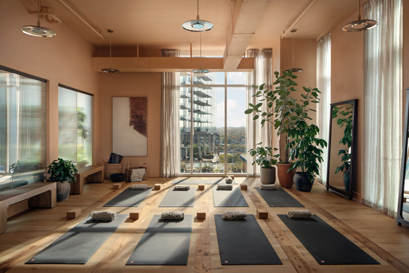 A modern yoga studio at Roost Rainey with large windows, potted plants, neatly arranged yoga mats, blocks, and bolsters on a wooden floor, offering city building views outside.