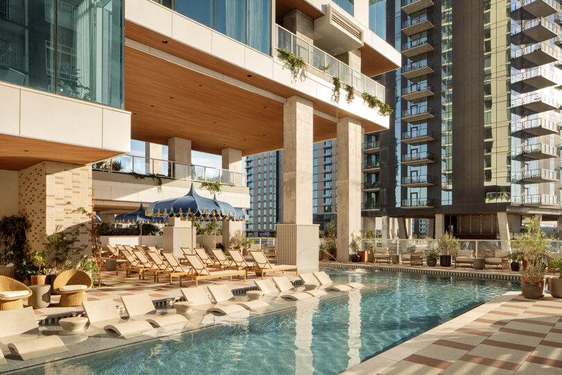 Modern outdoor pool area at Roost Rainey featuring submerged lounge chairs, stylish wooden deck seating, and tall glass buildings as the urban backdrop.