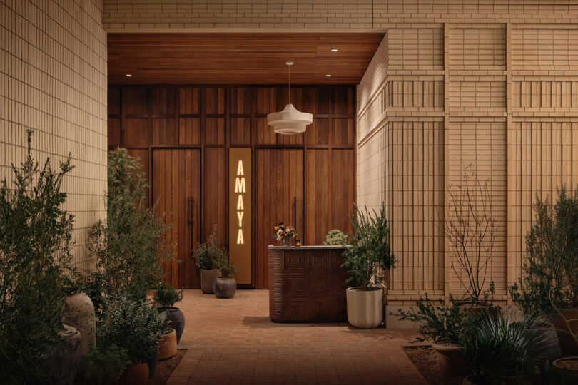 Entrance to Roost Rainey features wooden doors, a lit vertical "AMAYA" sign, potted plants, and a reception desk in a modern, neutral-toned setting.