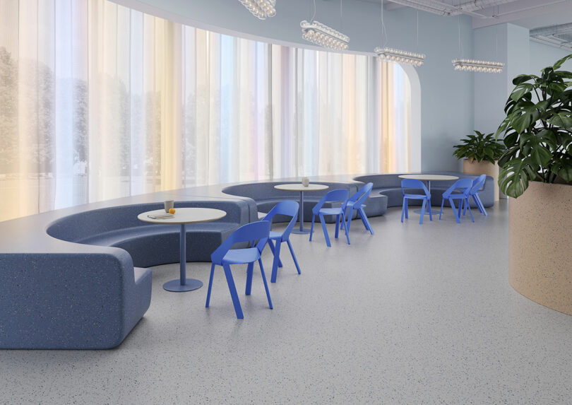 Modern lounge area with curved blue sofas, round tables, blue chairs, a large potted plant, and floor-to-ceiling windows with sheer, colorful curtains set atop elegant Tarkett flooring.