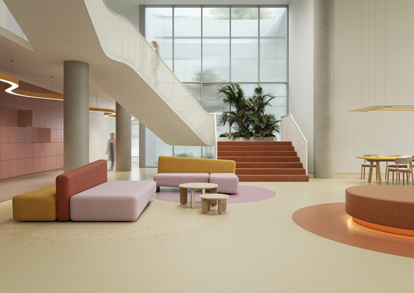 Modern, airy lobby with pastel seating, round tables, a stunning Tarkett terrazzo floor, a staircase, large windows, and indoor plants. A blurred figure is walking in the background.