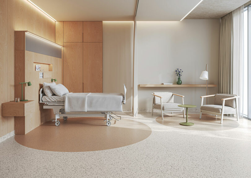 Modern hospital room with a single bed, Tarkett flooring, wooden cabinetry, two chairs, a small table, and a large window filling the space with natural light.