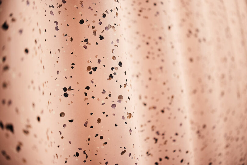Close-up of a light pink Tarkett surface with scattered small black and brown specks, possibly a patterned fabric or wallpaper.