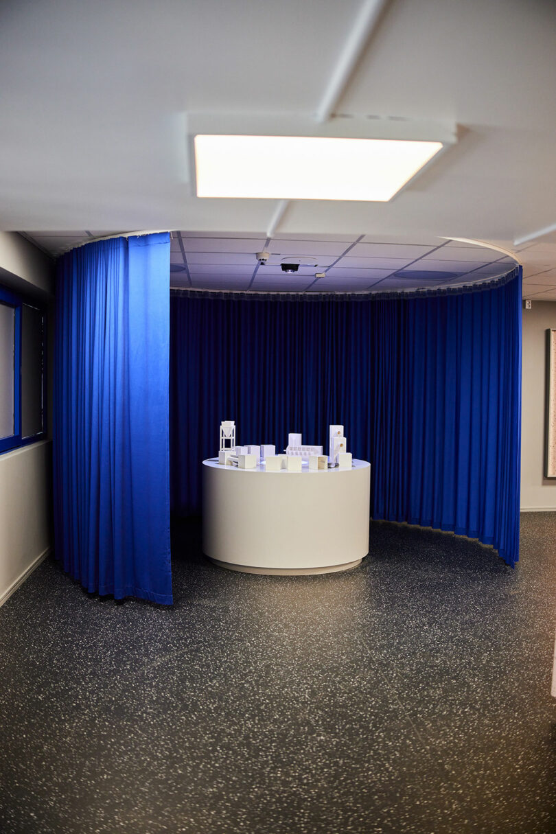 A round white display table with small white models sits in a room featuring blue curtains and sleek Tarkett speckled black flooring.