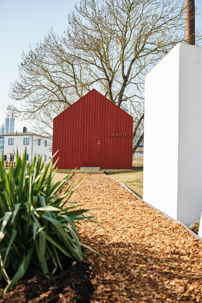 A wood chip pathway leads to a small red Tarkett building, nestled among grass and trees, with a white building visible in the background.