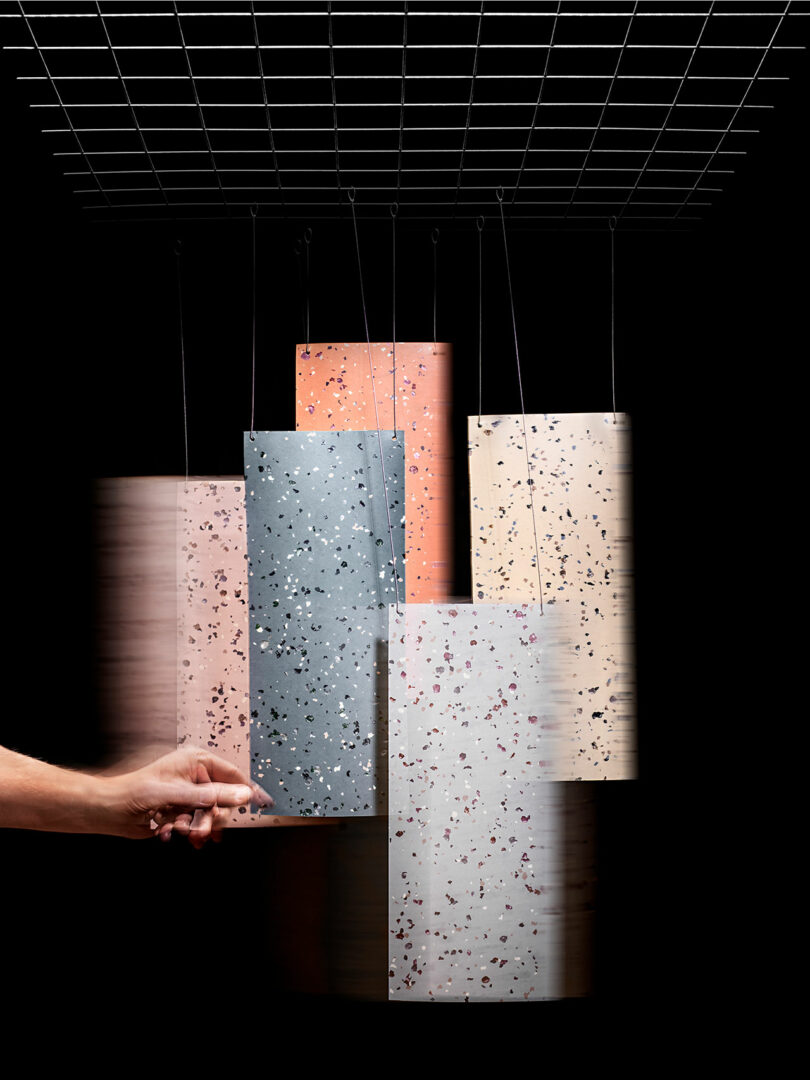 A hand moves one of five rectangular Tarkett terrazzo-like panels hanging from wires attached to a grid against a black background.