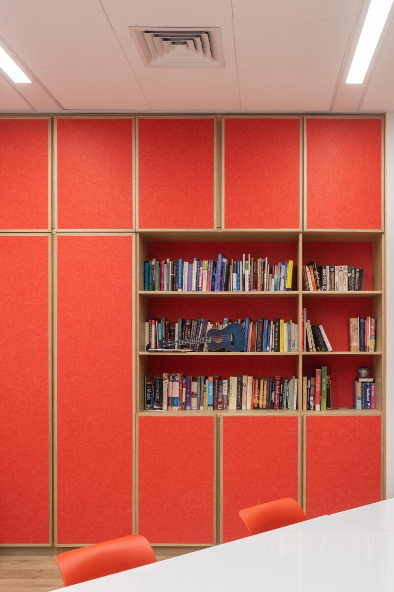 A built-in bookshelf with books is set in a wall with red panels above and below, next to a white table and orange chairs in a modern office or meeting room.