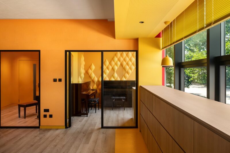 A modern room with orange and yellow walls, large windows with yellow blinds, and a small glass-enclosed area containing a piano and stool.