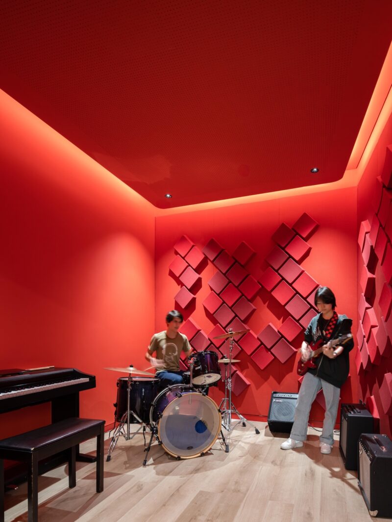 Two people play music in a bright red room with a drum set, electric guitar, keyboard, and sound-absorbing panels on the walls.