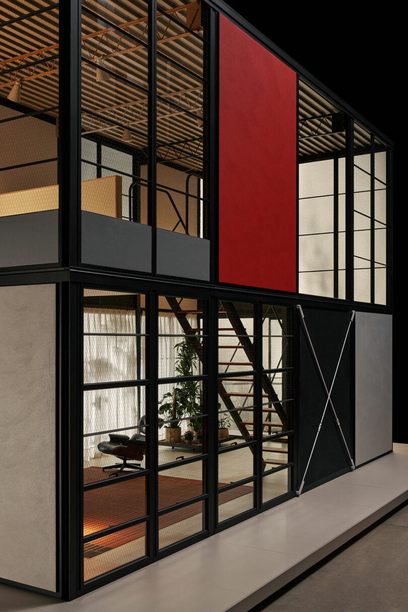 Two-story modern building with glass walls, black metal framing, a staircase, and red accent panel; interior features industrial decor and potted plants.