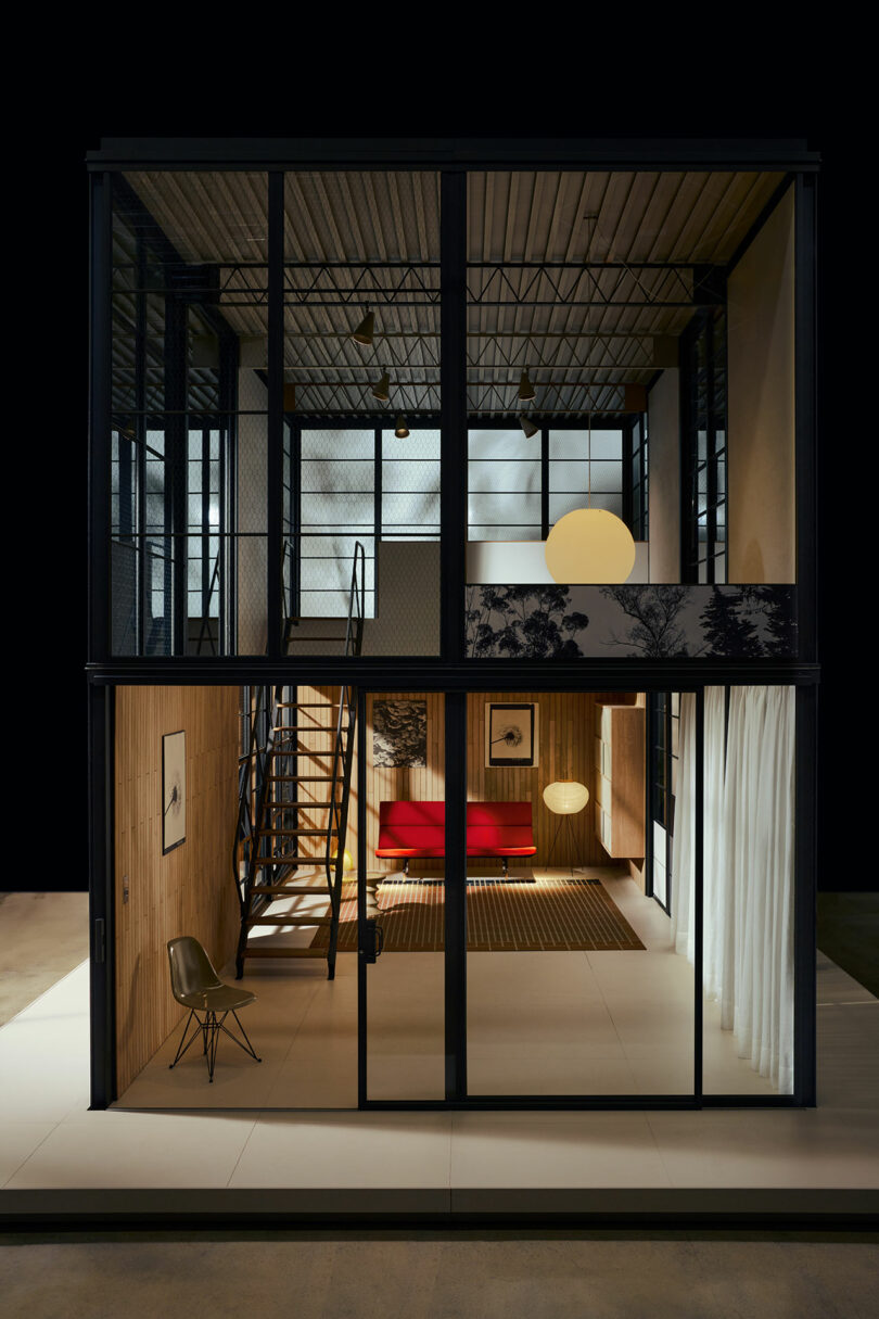 Two-story modern interior with large glass windows, exposed metal beams, wooden walls, a red sofa, spiral staircase, hanging globe lights, and minimal furnishings.