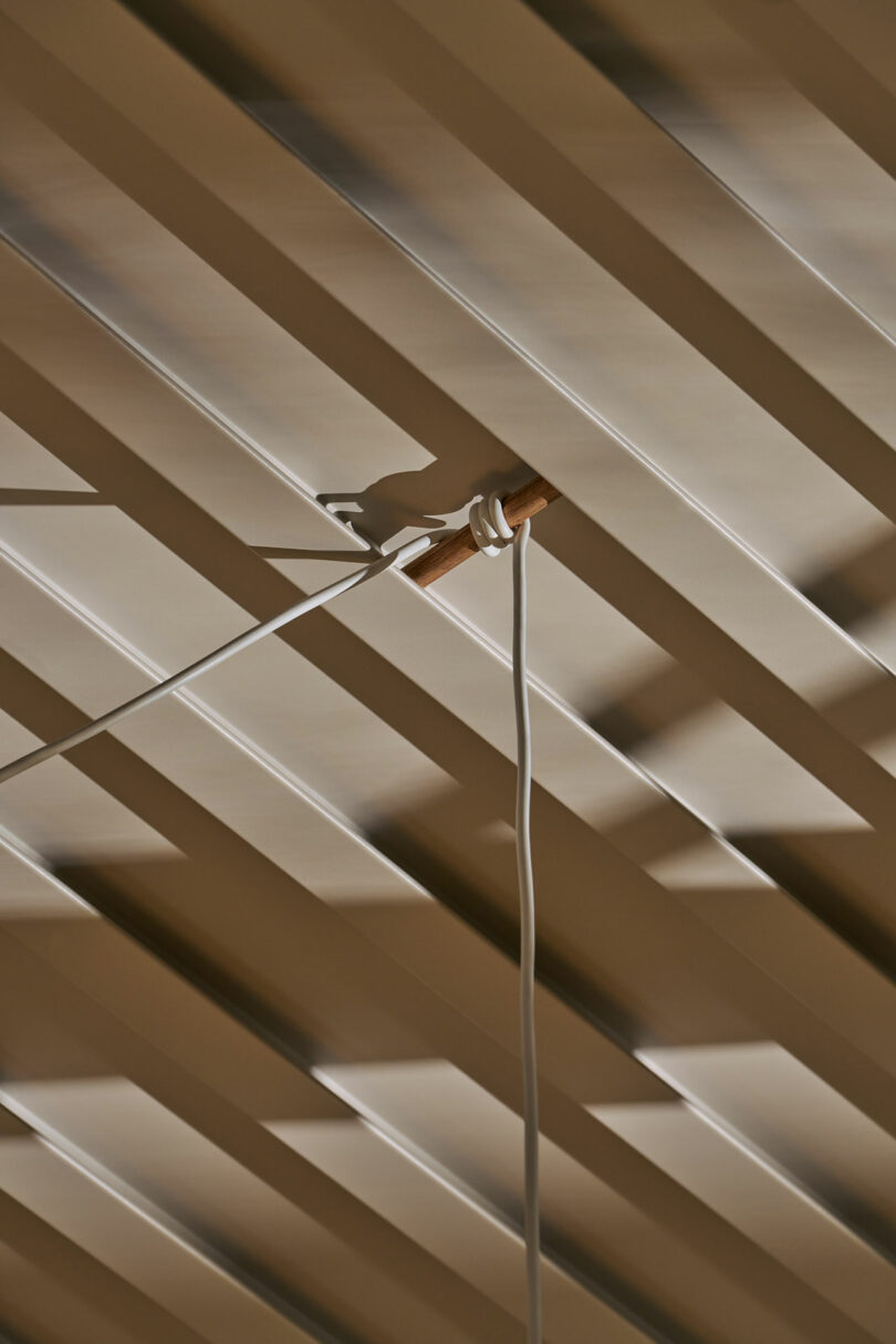 A white cord is tied around a wooden dowel and hangs from a ceiling with diagonal, shadowed slats.