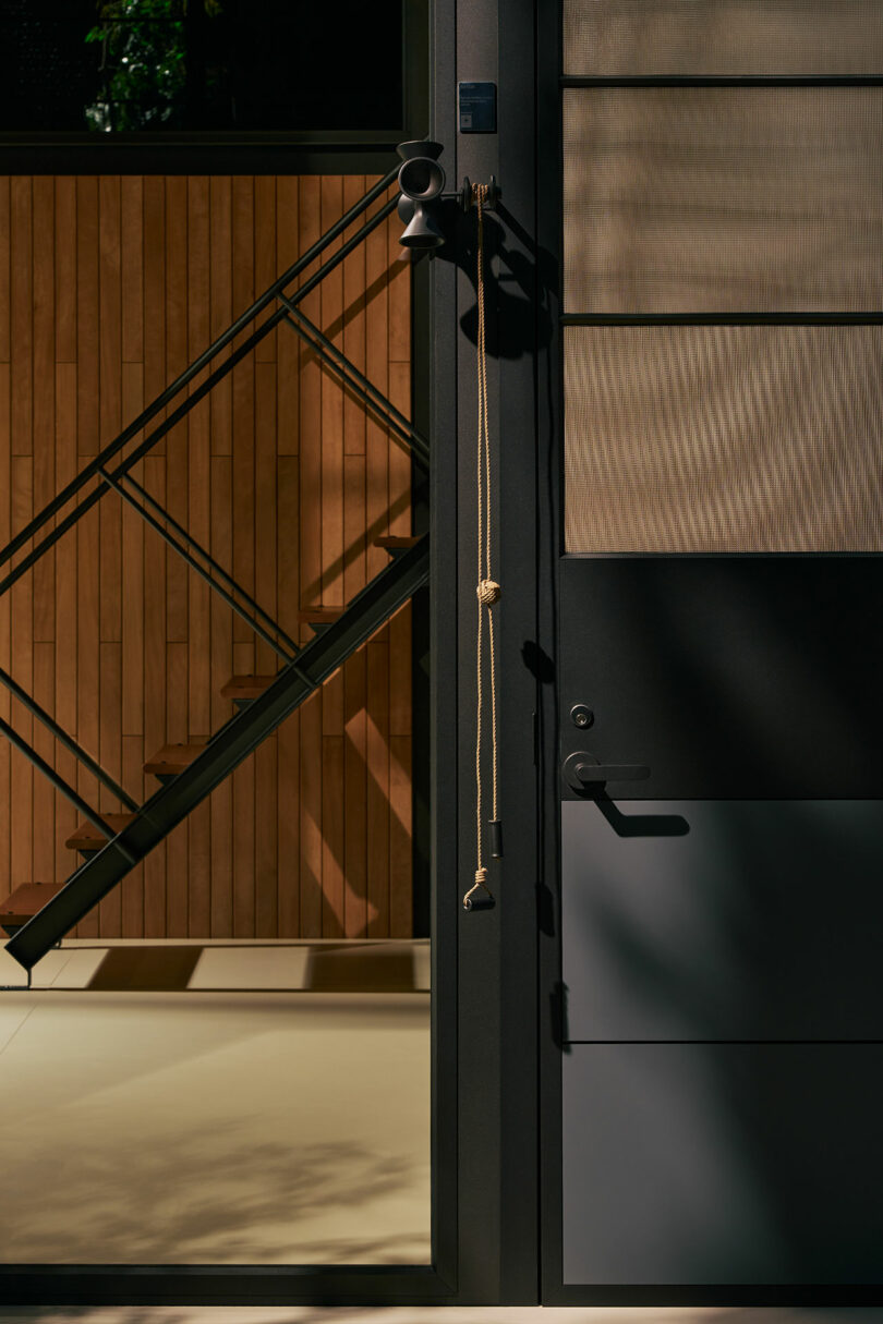 A modern black door is partially open, revealing an interior with wood paneling, a staircase, and a long cord with knots hanging from the door handle.