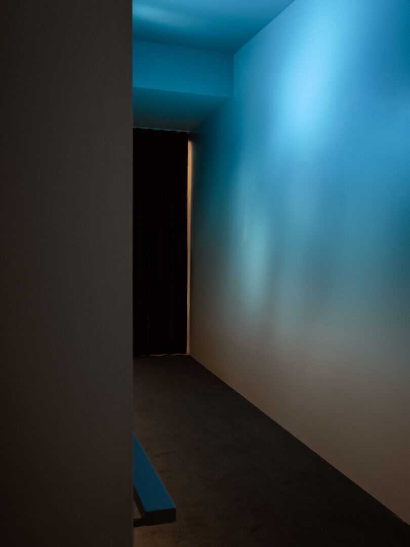 A narrow hallway with blue lighting leads to a dark, curtained doorway. Shadows and light create a calm, minimal atmosphere.