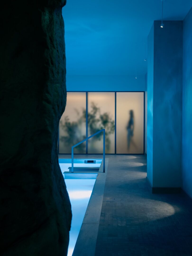 An indoor pool area with blue lighting, frosted glass panels, and the blurred silhouette of a person standing behind the glass.