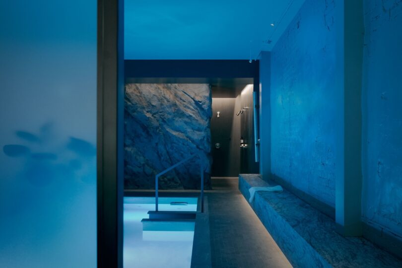 Modern indoor spa room with blue lighting, a small pool with steps, a stone-textured wall, and a bench with a towel on it.
