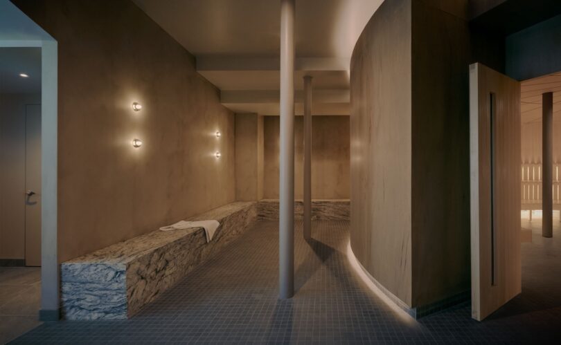 A minimalist spa interior with stone benches, wall-mounted lights, tiled floor, and curved wooden walls; a white towel is placed on one bench.
