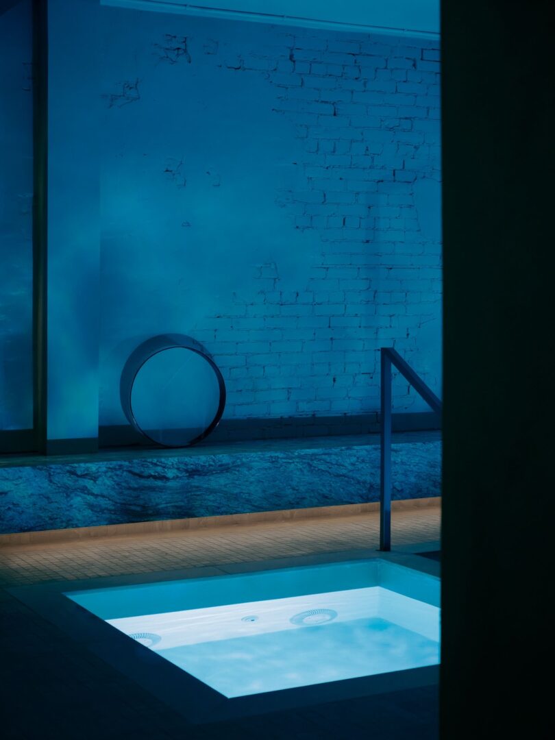 A small illuminated indoor pool with steps leading in, surrounded by blue lighting and a textured brick wall in the background.