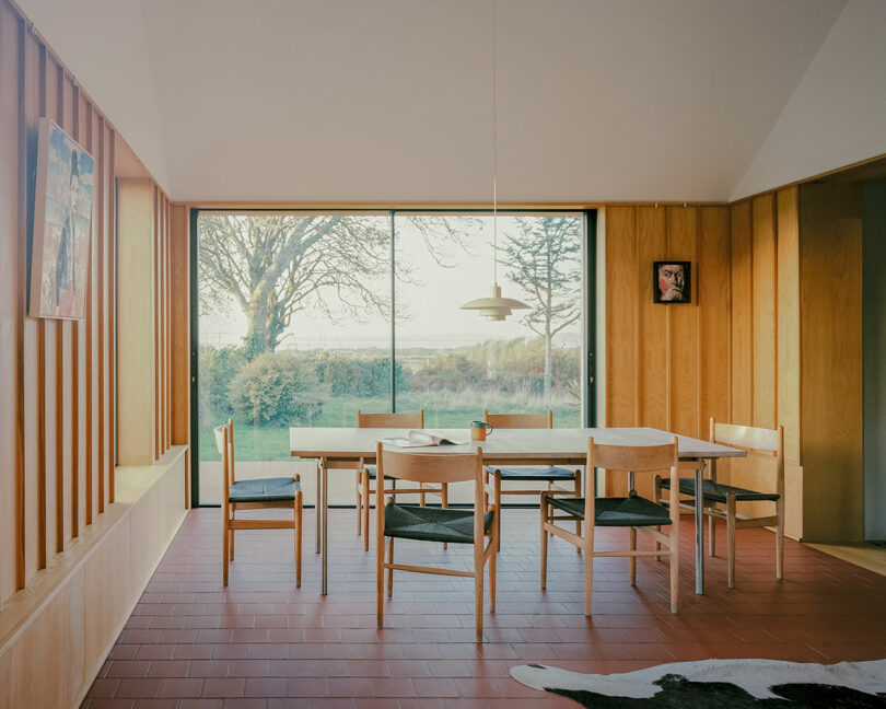 Minimalist dining room at Delfyd Farm with wooden walls, large glass sliding doors overlooking a garden, a rectangular table with six chairs, a pendant light above, and artwork on the walls.