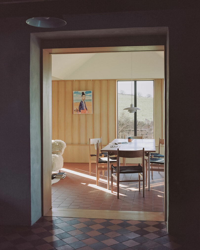 A dining area with wooden walls, a table with chairs, a pendant light, a window with a scenic view, and a portrait painting on the wall.