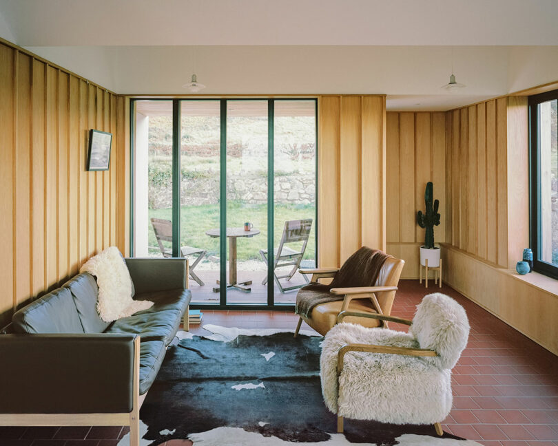 Modern living room with wood-paneled walls, large windows, two chairs, a sofa with sheepskin throws, a cowhide rug, and a cactus in a white planter. Patio set visible outside.