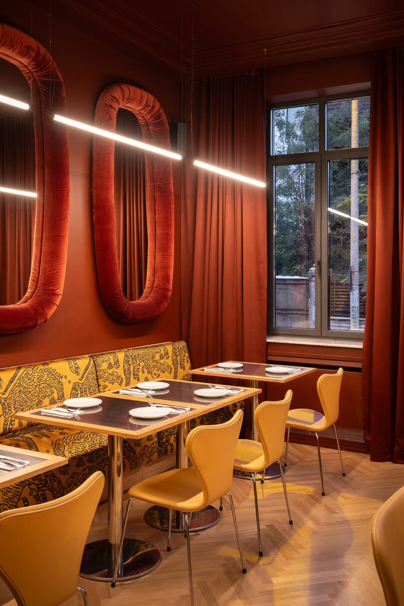 Modern restaurant interior with orange walls, large red velvet mirrors, yellow chairs, patterned bench seating, set tables, and a window showing trees outside.