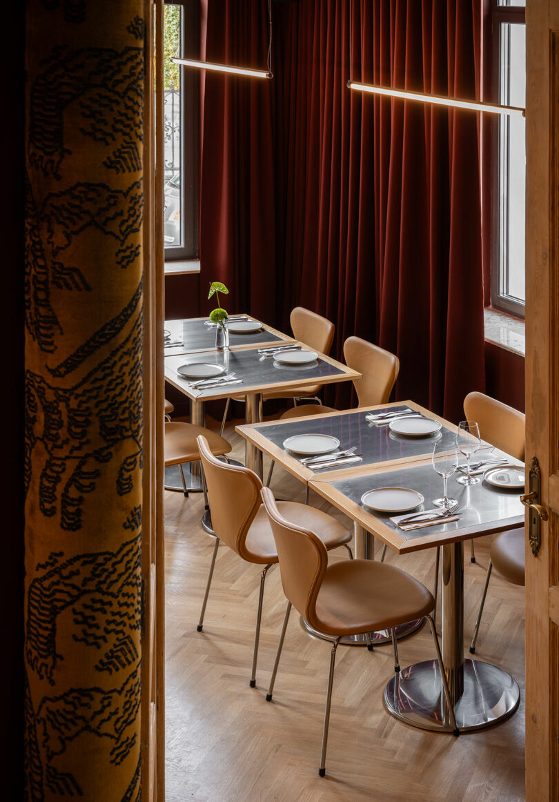 A small restaurant dining area with tan chairs, metal tables set with plates and glasses, wooden floors, and red curtains covering the windows.