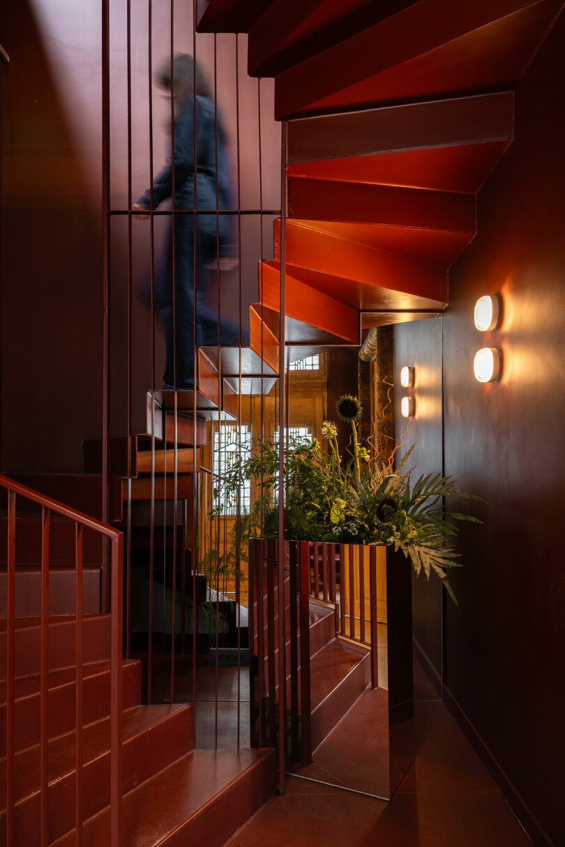 A person walks upstairs on a red spiral staircase in a dimly lit interior with plants and round wall lights.