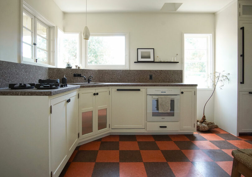 Modern kitchen with white cabinets, brown speckled countertops, gas stove, built-in oven, and orange-and-brown checkered floor tiles; natural light from two windows.