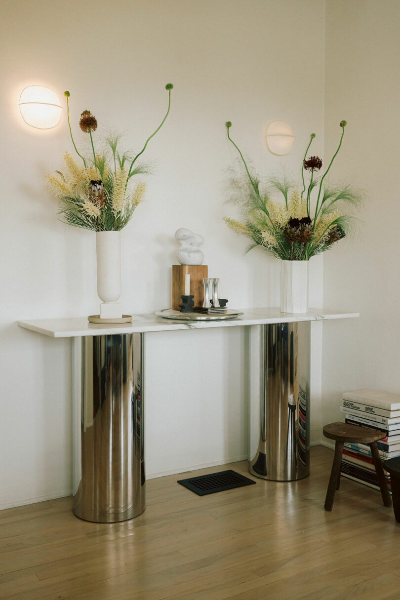 A minimalist console table with metallic cylindrical legs, decorated with tall floral arrangements, sculptural decor, and books stacked on a stool beside it.