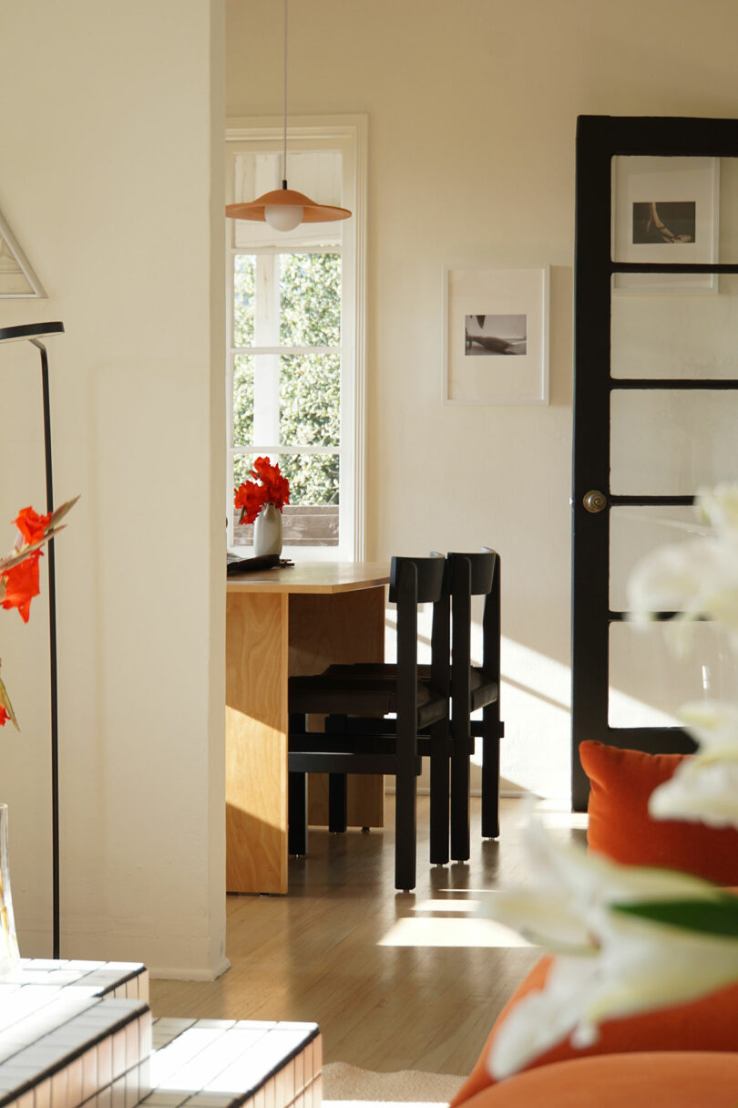 A small dining table with two black chairs is set near a window with sunlight streaming in. A vase with red flowers sits on the table in a bright, modern living space.