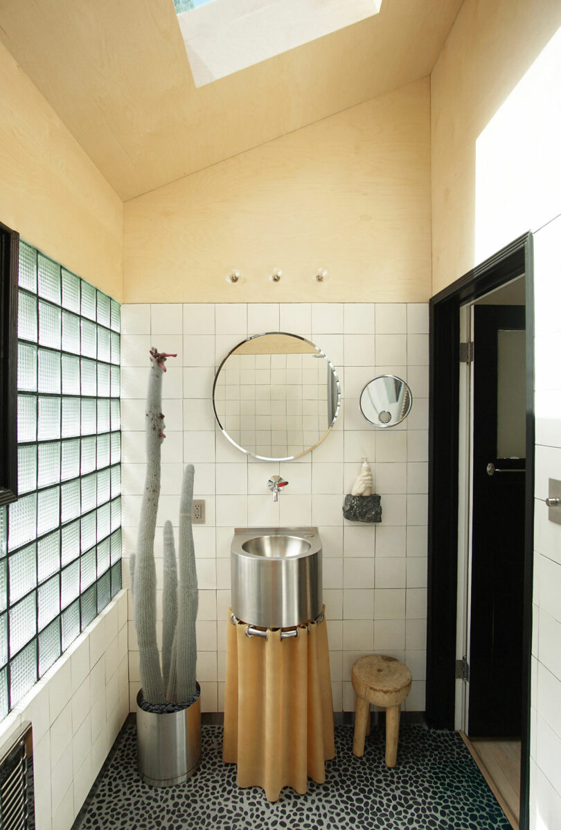 A small bathroom with white tiled walls, a round mirror above a stainless steel sink, a cactus, a wooden stool, and pebble-patterned floor, lit by a skylight.