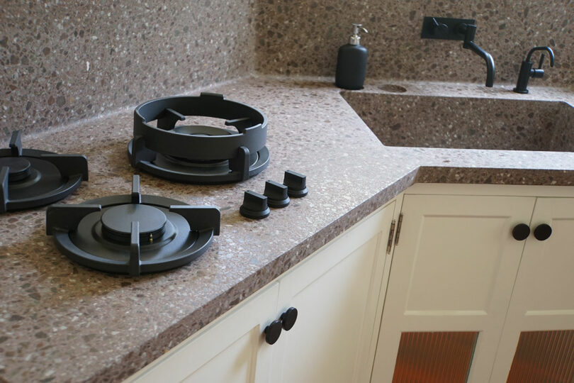 A modern kitchen counter with two gas burners, burner caps, a soap dispenser, a sink with a black faucet, and white cabinets with black handles.