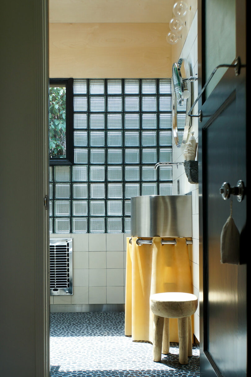 Modern bathroom with glass block window, stainless steel sink, round mirror, yellow fabric sink skirt, small wooden stool, and pebble-patterned floor.