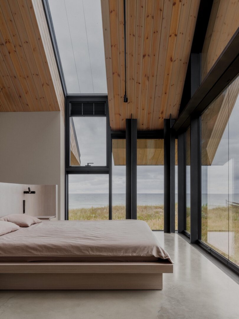 Minimalist bedroom with a platform bed, wood-paneled ceiling, large floor-to-ceiling windows, and a view of grassy dunes and the sea under a cloudy sky.