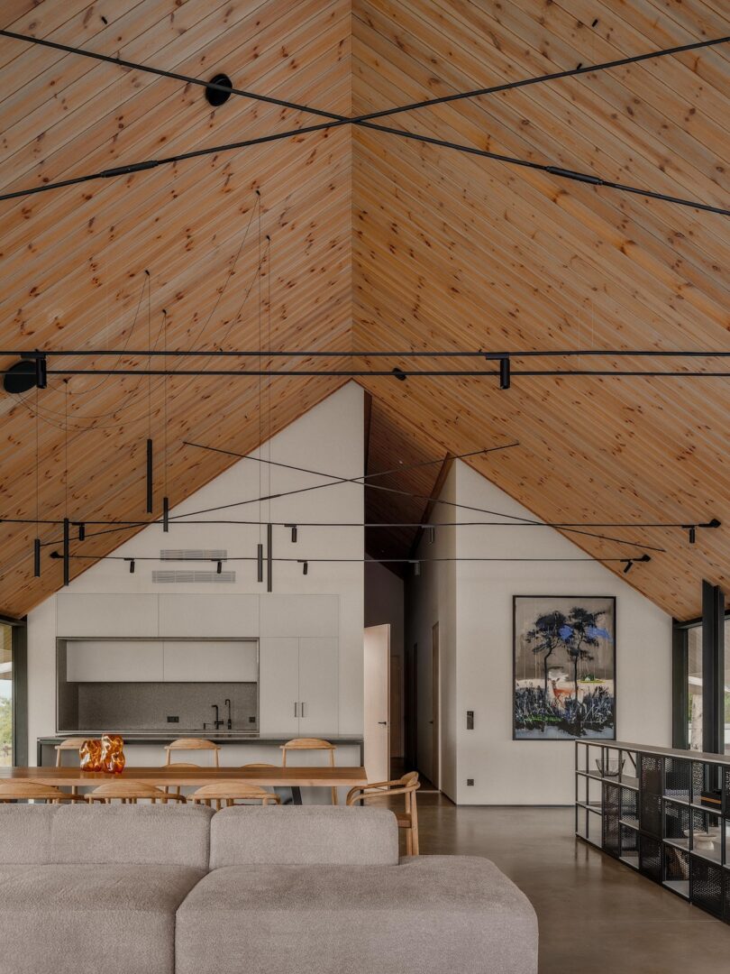 Modern open-plan kitchen and dining area with sloped wooden ceiling, minimalist white cabinetry, pendant lighting, and wall art.